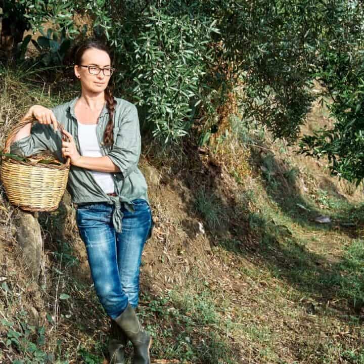 A woman wearing glasses, jeans, and boots leans against a slope while holding a basket in an outdoor, rural setting with trees and grass.