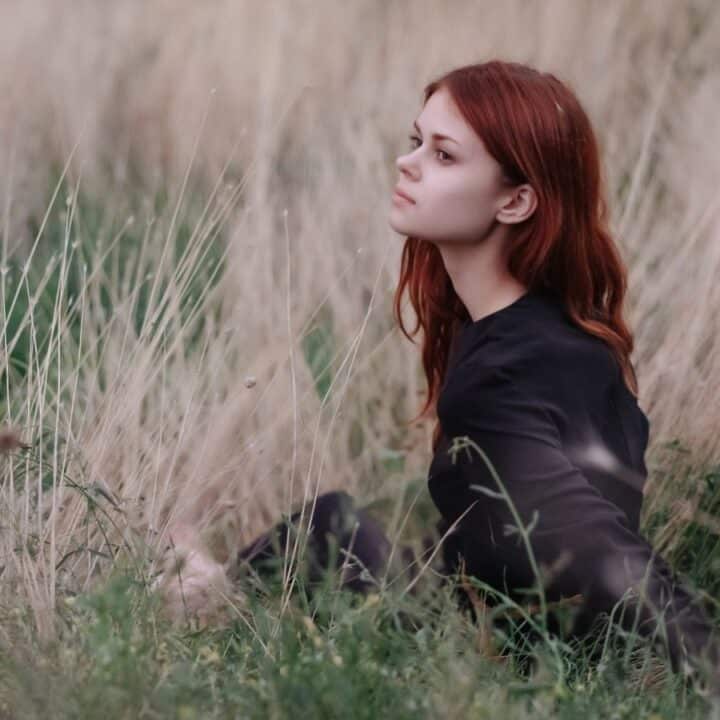 A person with long red hair, wearing a black top, sits in tall dry grass and green foliage, looking off to the side.