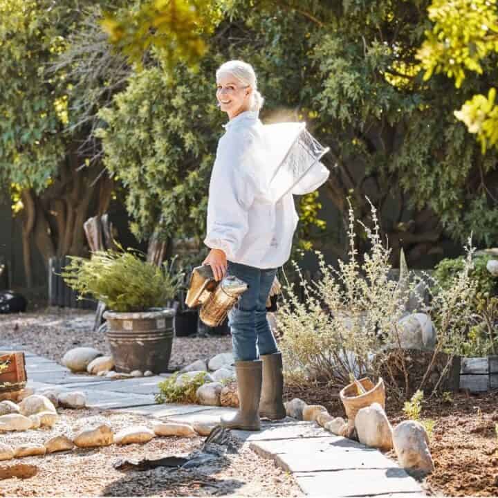 A person wearing a white shirt, jeans, and boots stands outdoors on a stone path in a garden, holding gardening tools and looking back over their shoulder.