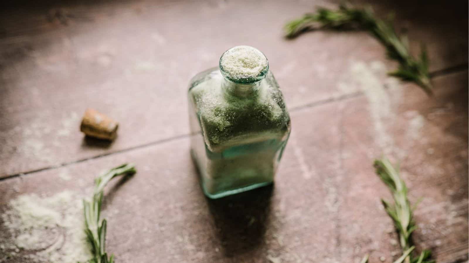 A glass bottle filled with coarse green salt sits on a wooden surface, surrounded by scattered rosemary sprigs and a cork.