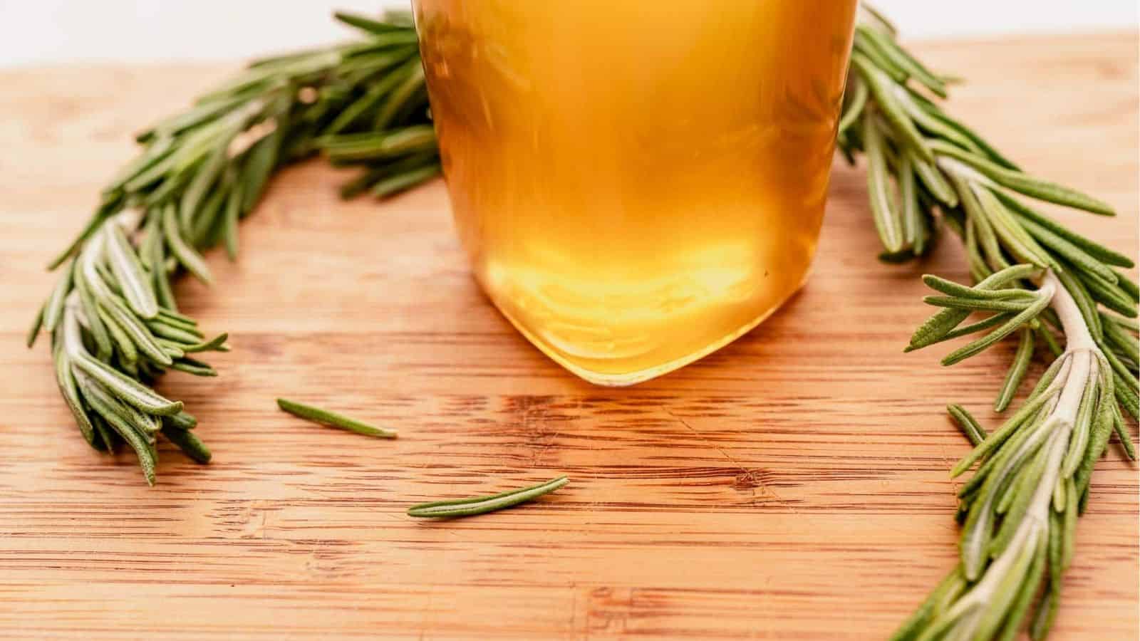 A glass of golden liquid placed on a wooden surface, surrounded by a semicircle of fresh rosemary sprigs.