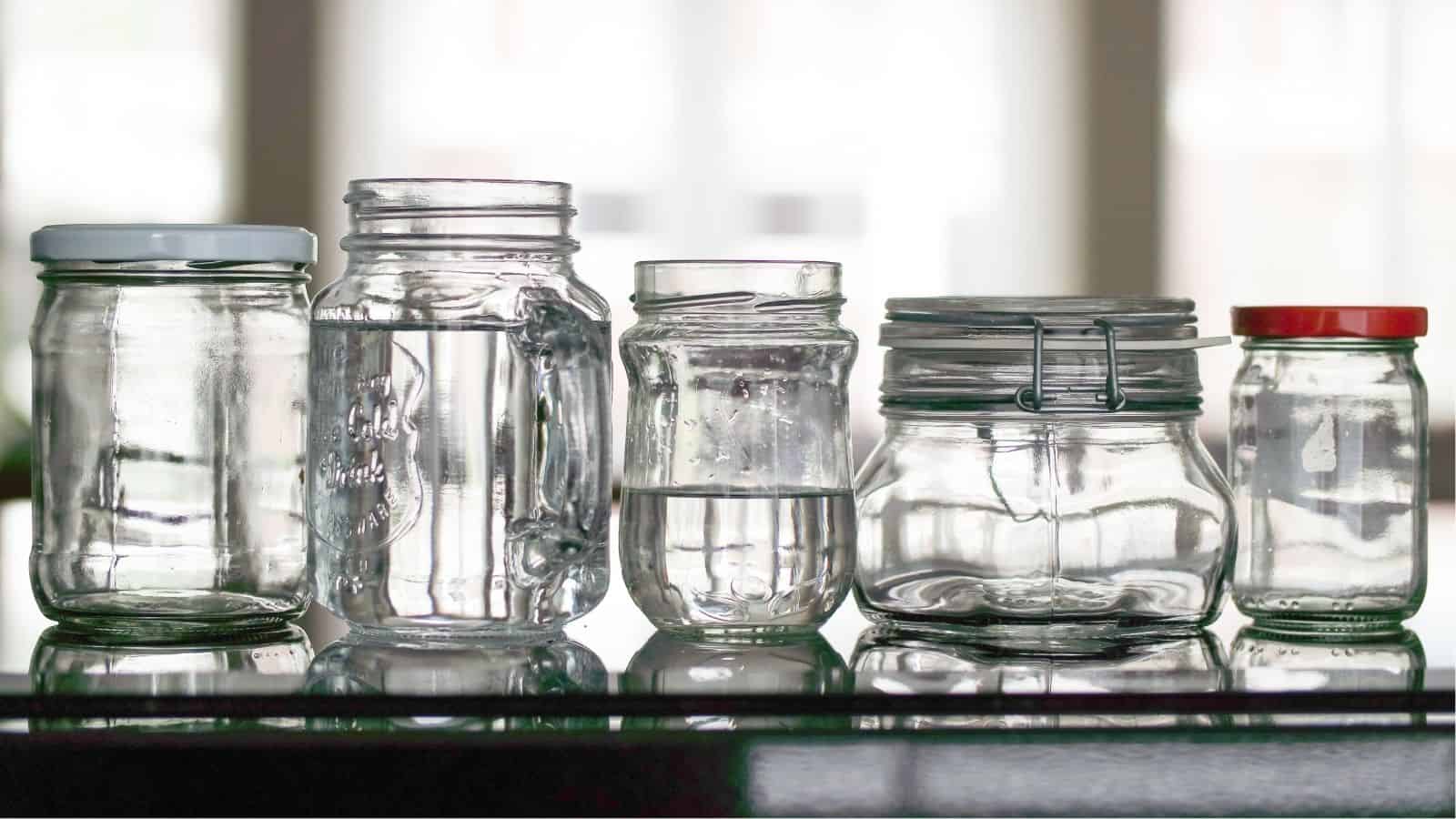 Five glass jars of various sizes and shapes, some with lids, are placed side by side on a reflective surface.