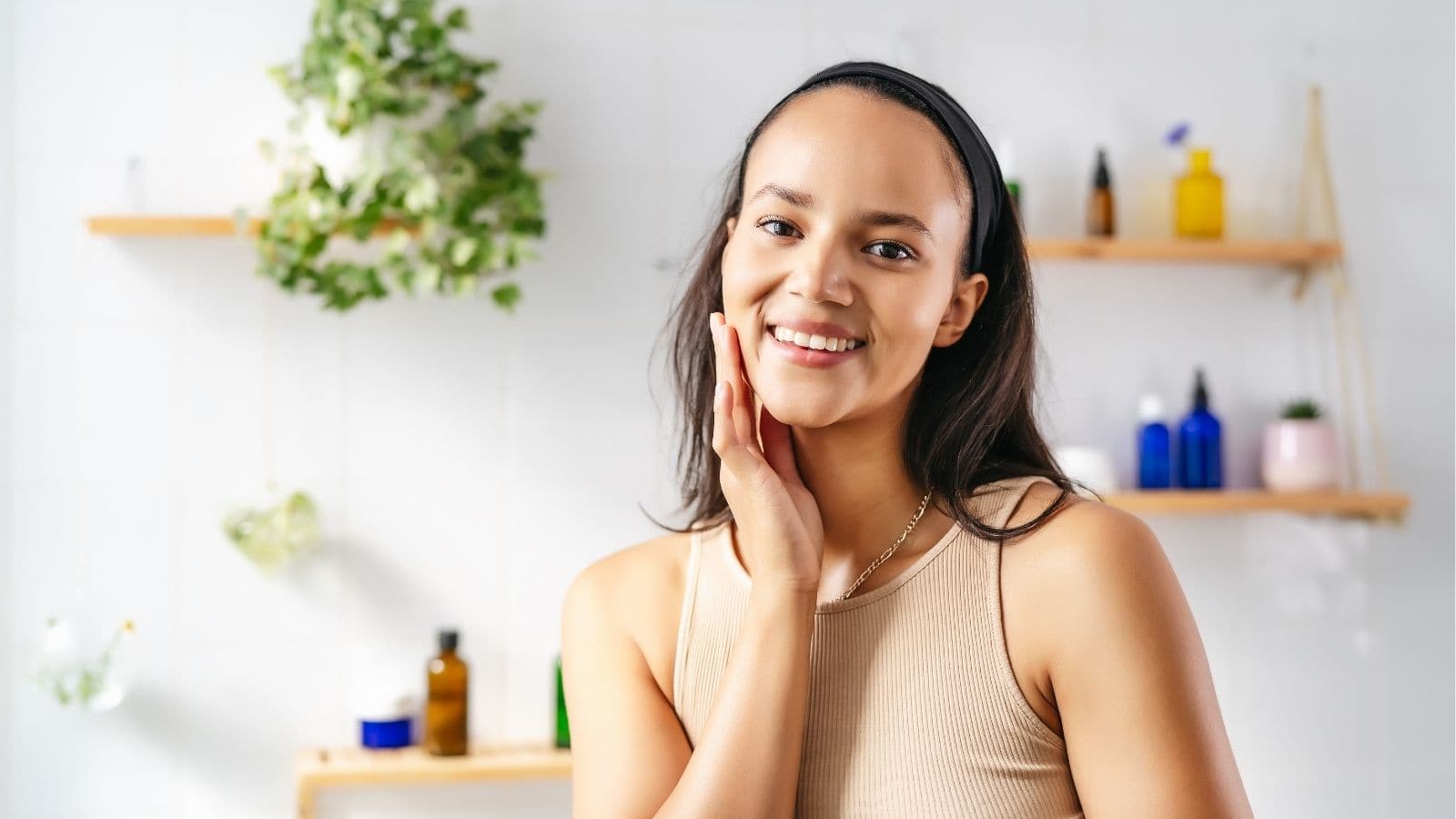 A woman with long dark hair, wearing a beige tank top and black headband, smiles and touches her face in a bathroom with plants and skincare bottles on shelves in the background.