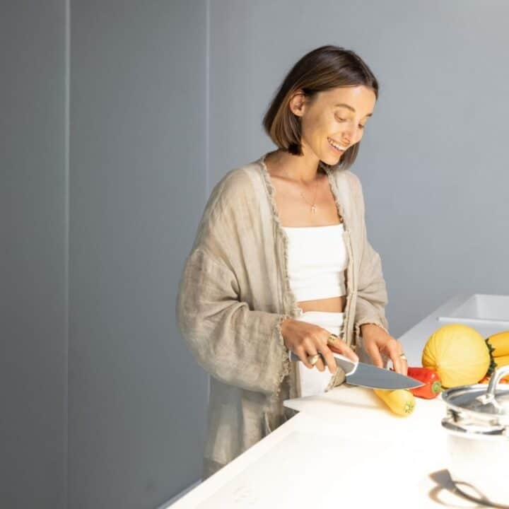 A woman in a light robe slices yellow squash on a kitchen counter with various vegetables and a pot nearby.