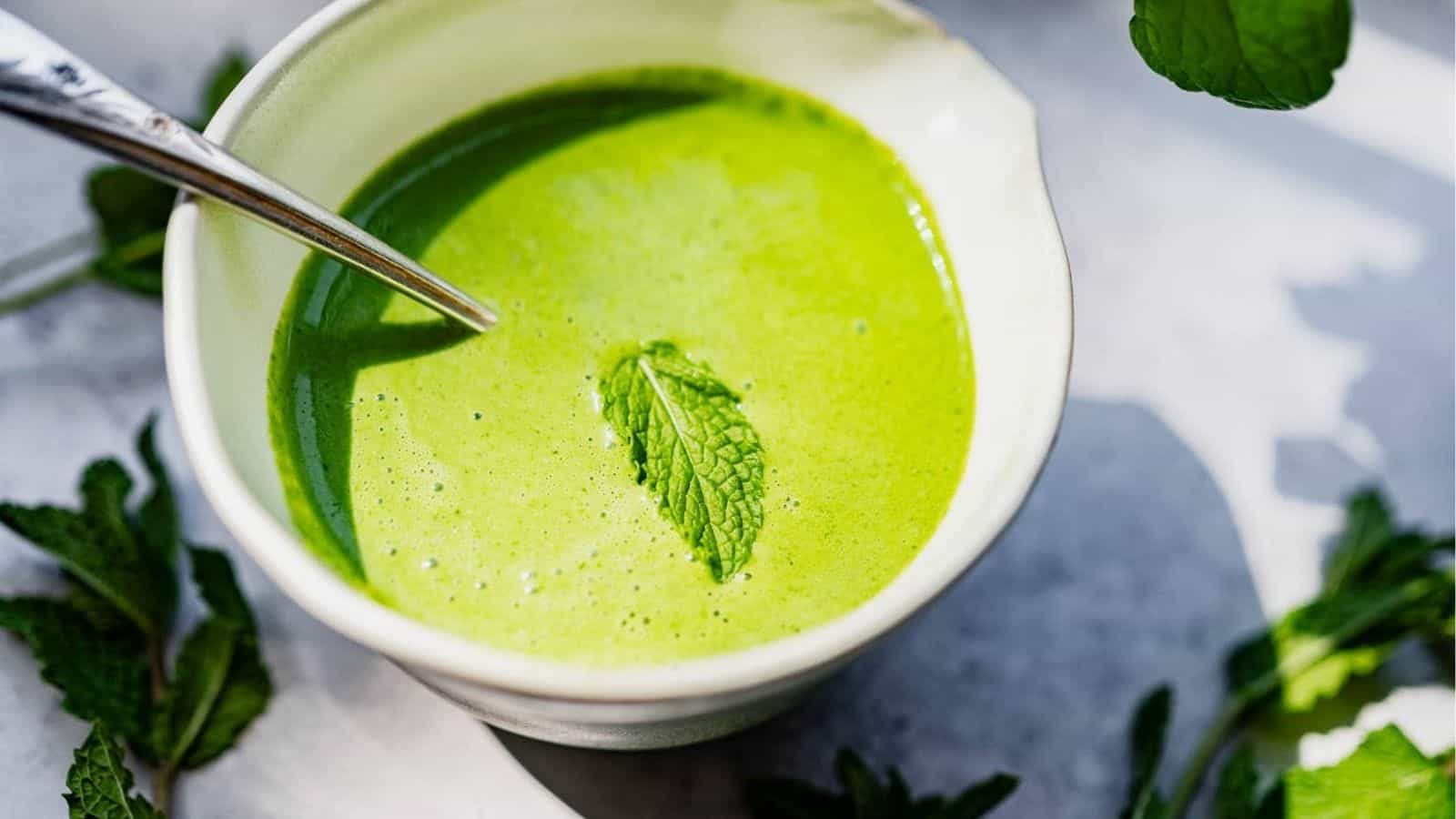A white bowl filled with green soup garnished with a mint leaf, with a spoon resting inside and mint leaves scattered nearby.