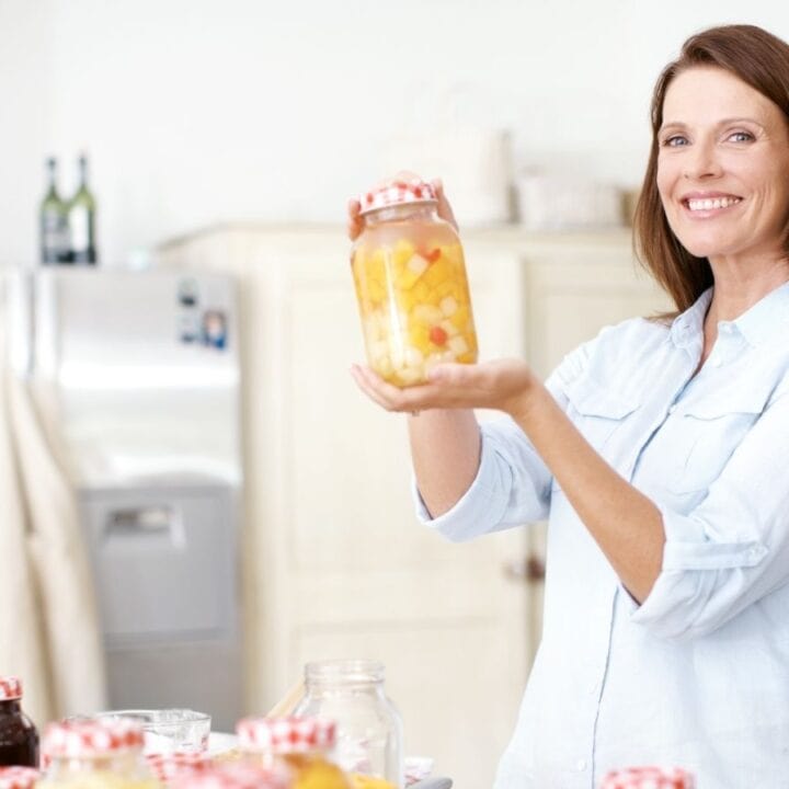 A woman in a kitchen holds up a jar of preserved fruit and smiles, with other similar jars on the counter in front of her.