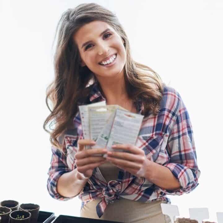 Woman in a plaid shirt stands indoors holding seed packets, smiling, with small plant pots and gardening supplies on the table in front of her.