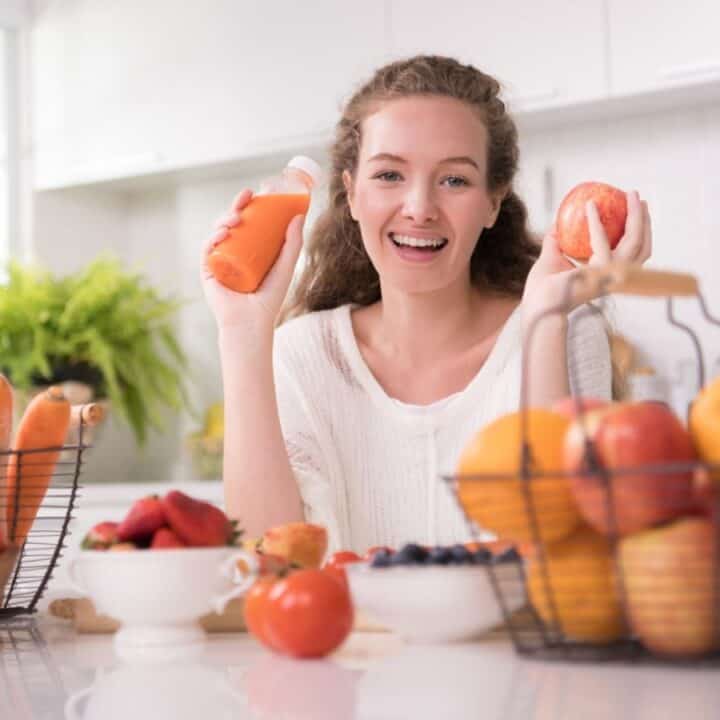 A woman in a kitchen smiles while holding a bottle of orange juice and an apple, surrounded by baskets and bowls of fresh fruit.