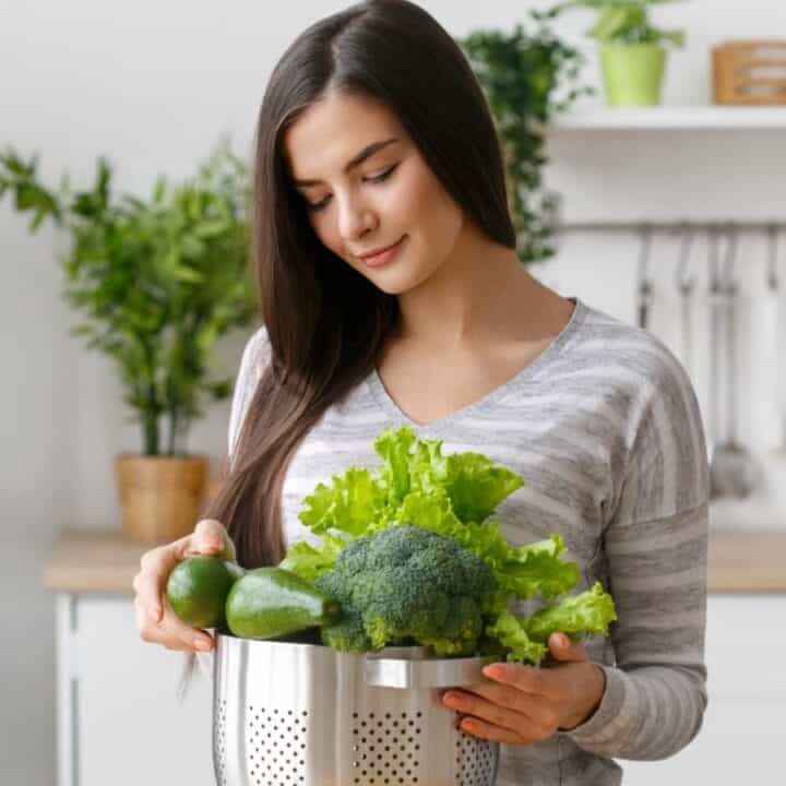 A woman in a kitchen holds a colander filled with fresh green vegetables, including lettuce, broccoli, and cucumbers.