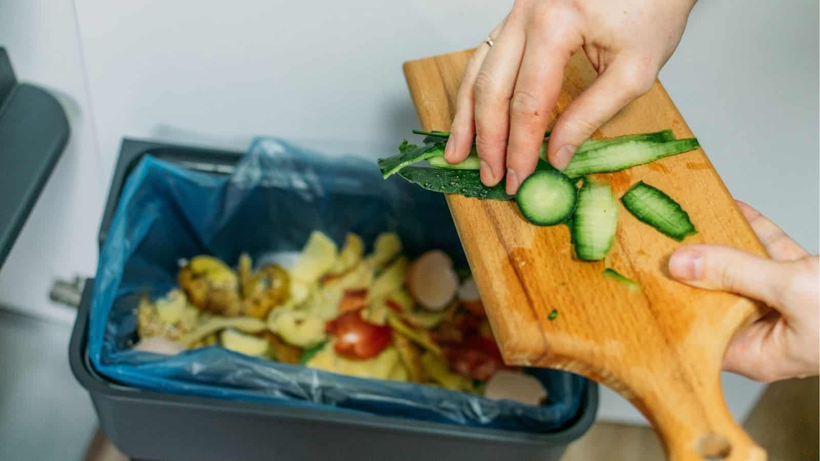 A person scraping vegetable scraps from a cutting board into a kitchen compost bin lined with a blue bag.