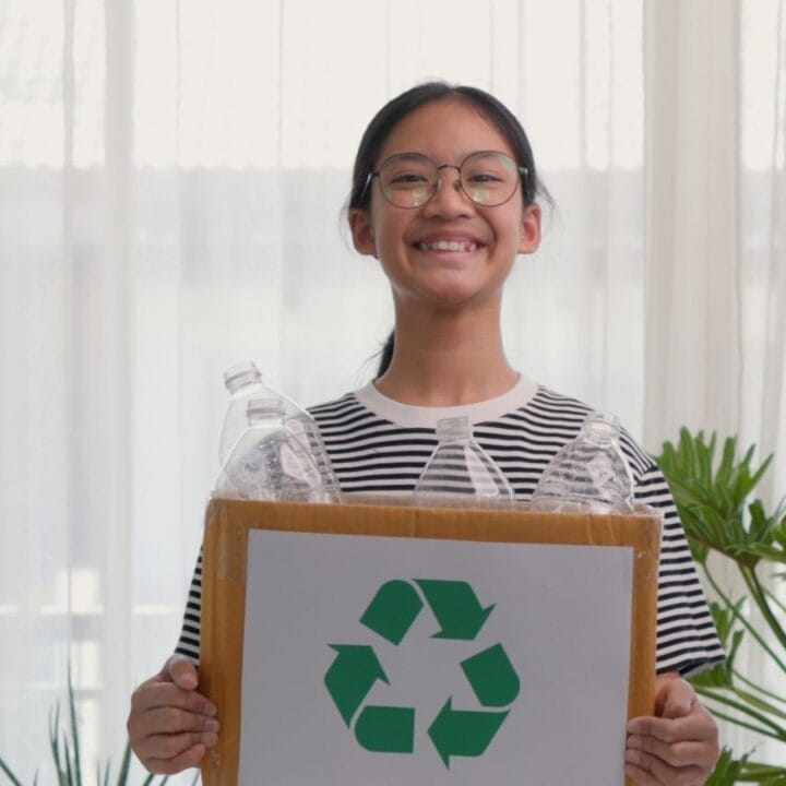 A person in glasses and a striped shirt holds a box with a recycling symbol and plastic bottles, standing indoors among green plants.