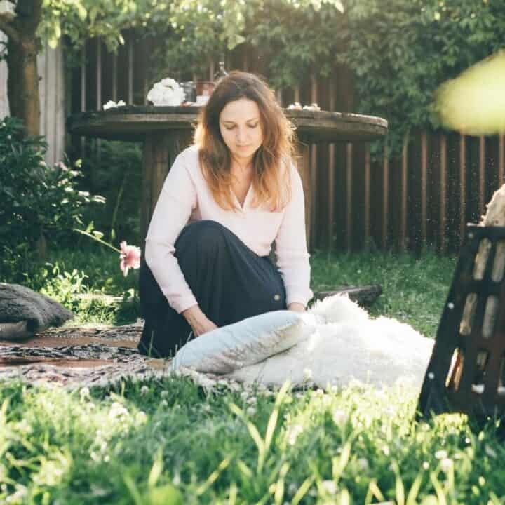 A woman sits on cushions and a patterned rug in a sunny garden, surrounded by flowers and greenery, with a wooden table and chairs in the background.