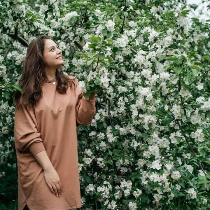 A woman in a brown dress stands outdoors next to a blooming tree with white flowers, looking slightly upward and smiling.