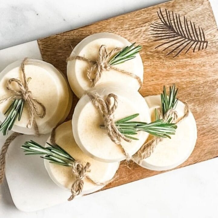 Six round, white soap bars tied with twine and sprigs of rosemary are arranged on a wooden and white cutting board with a leaf design, set on a marble surface.