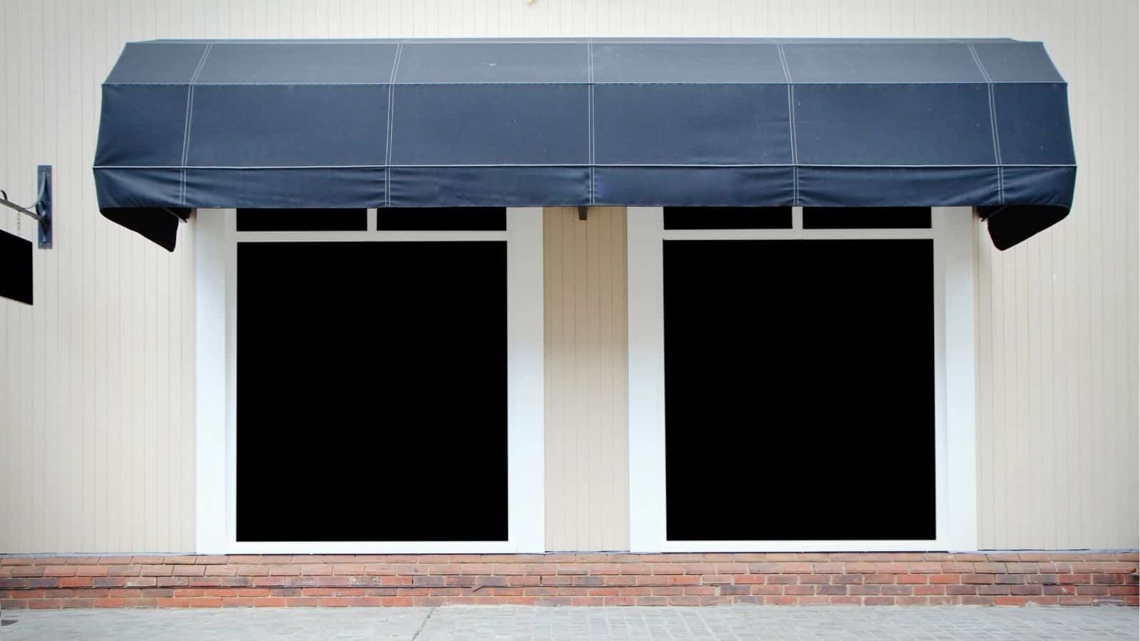 Two large, empty storefront windows with white frames and a black awning above, set in a beige wall with a brick base.