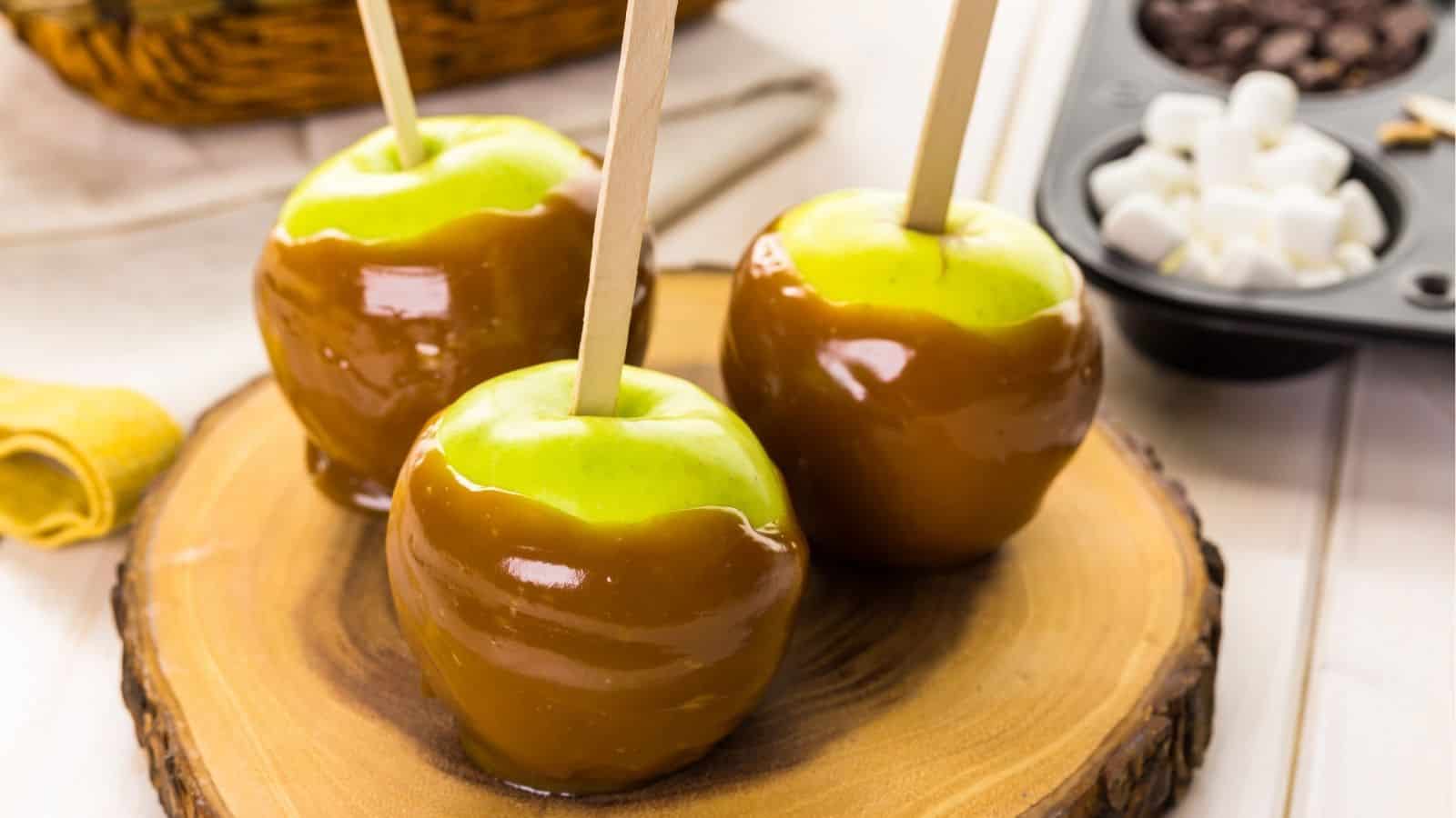 Three green apples on wooden sticks are coated in caramel and placed on a round wooden board, with marshmallows and chocolate chips in the background.
