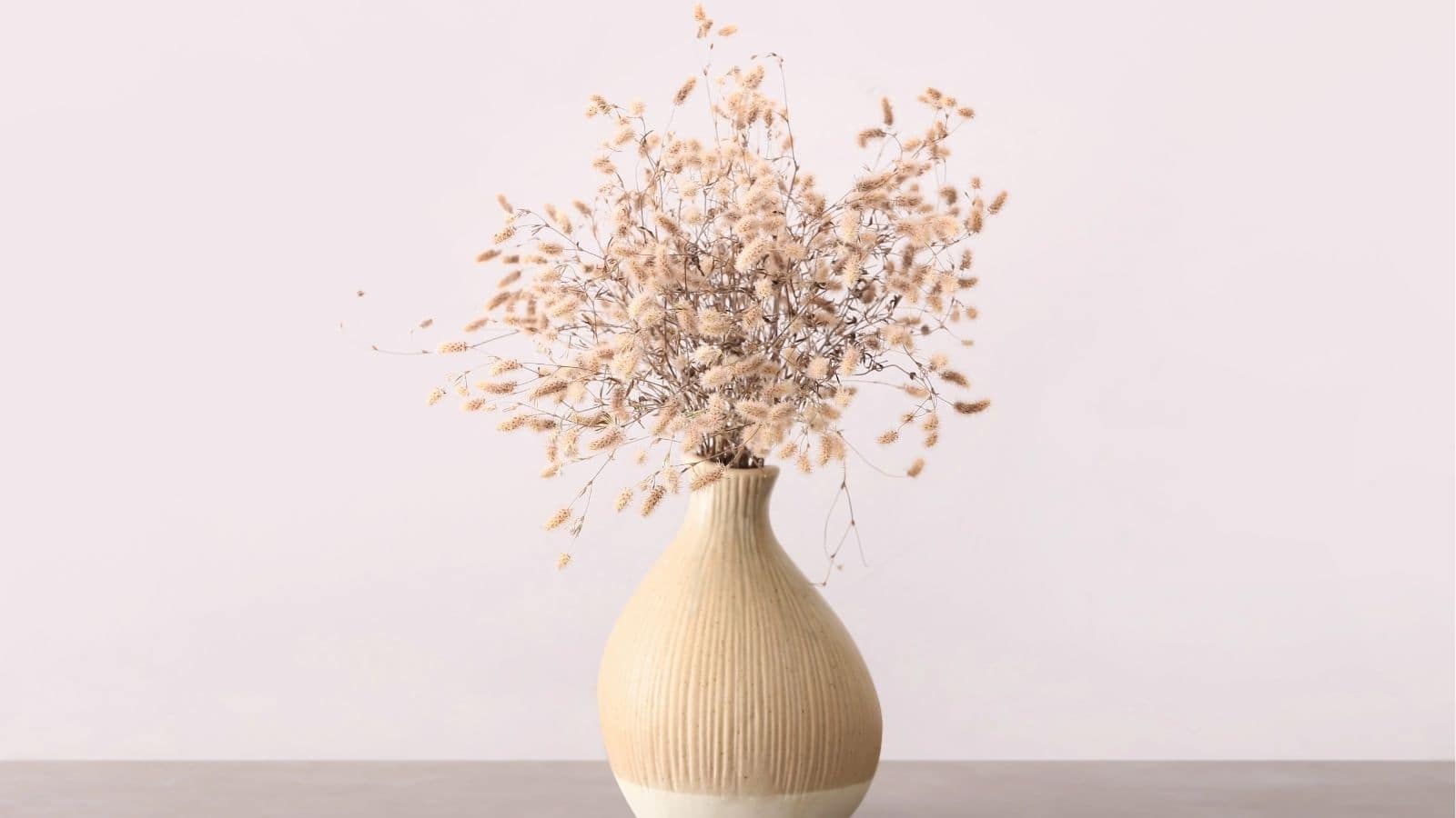 A beige ceramic vase holds a bundle of dried, light brown grasses, set against a plain, pale pink background.
