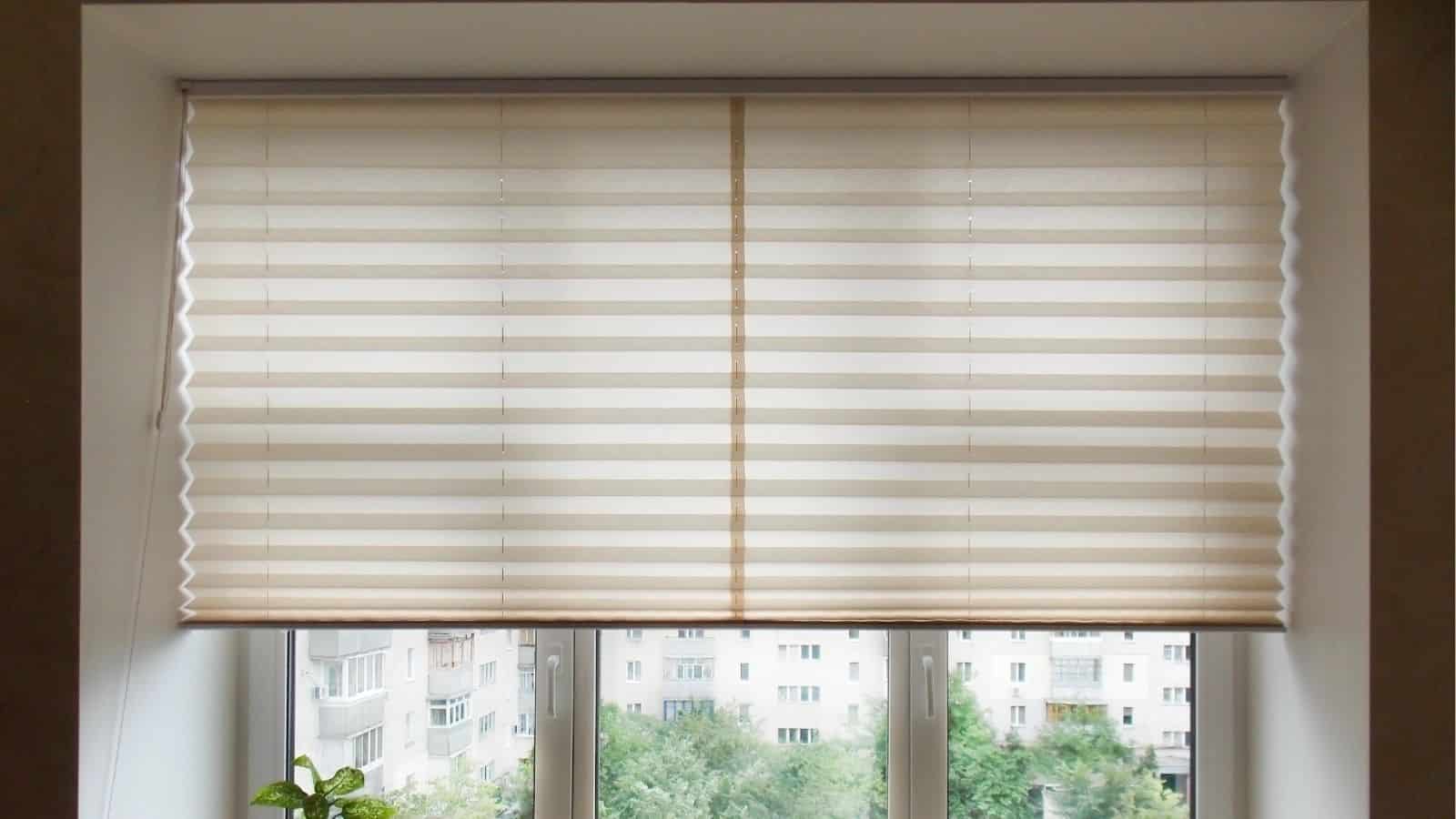 A window with sheer, horizontal striped blinds partially closed, overlooking an urban apartment building and greenery outdoors.