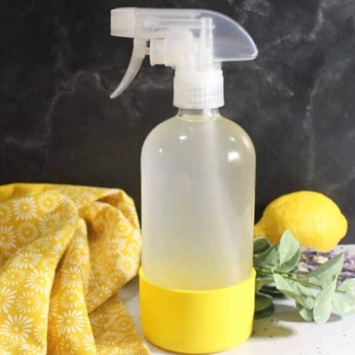 A clear spray bottle with a yellow base stands on a counter next to a yellow patterned cloth, a lemon, some greenery, and a sprig of lavender, against a dark background.