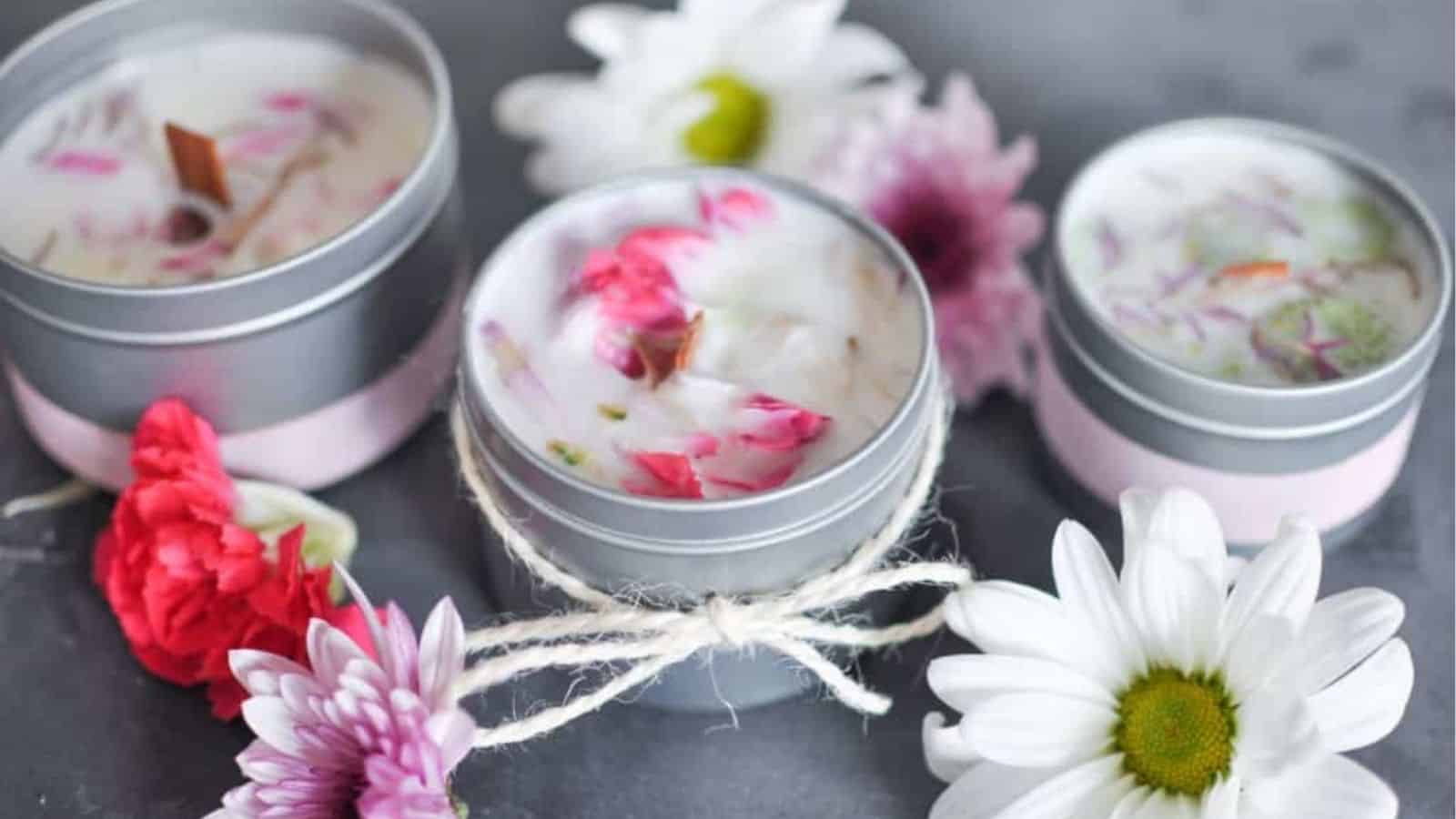Three small tin candles with flower petals in the wax are arranged on a surface, surrounded by white and pink daisies.