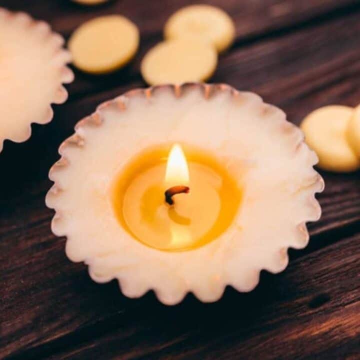 A lit candle in a fluted holder sits on a wooden surface, with another candle and scattered wax discs visible in the background.