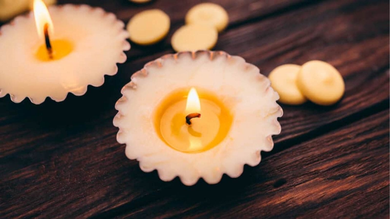 A lit candle in a fluted holder sits on a wooden surface, with another candle and scattered wax discs visible in the background.