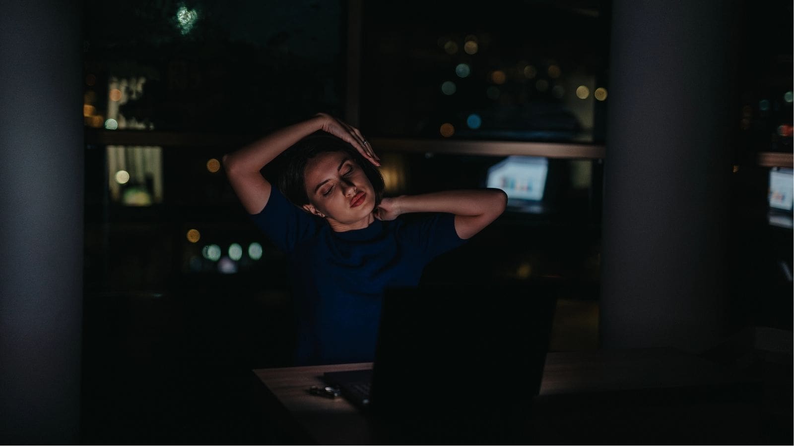A person sits at a desk in a dimly lit room, stretching their neck and arm while facing a laptop, with city lights visible through the window behind them.