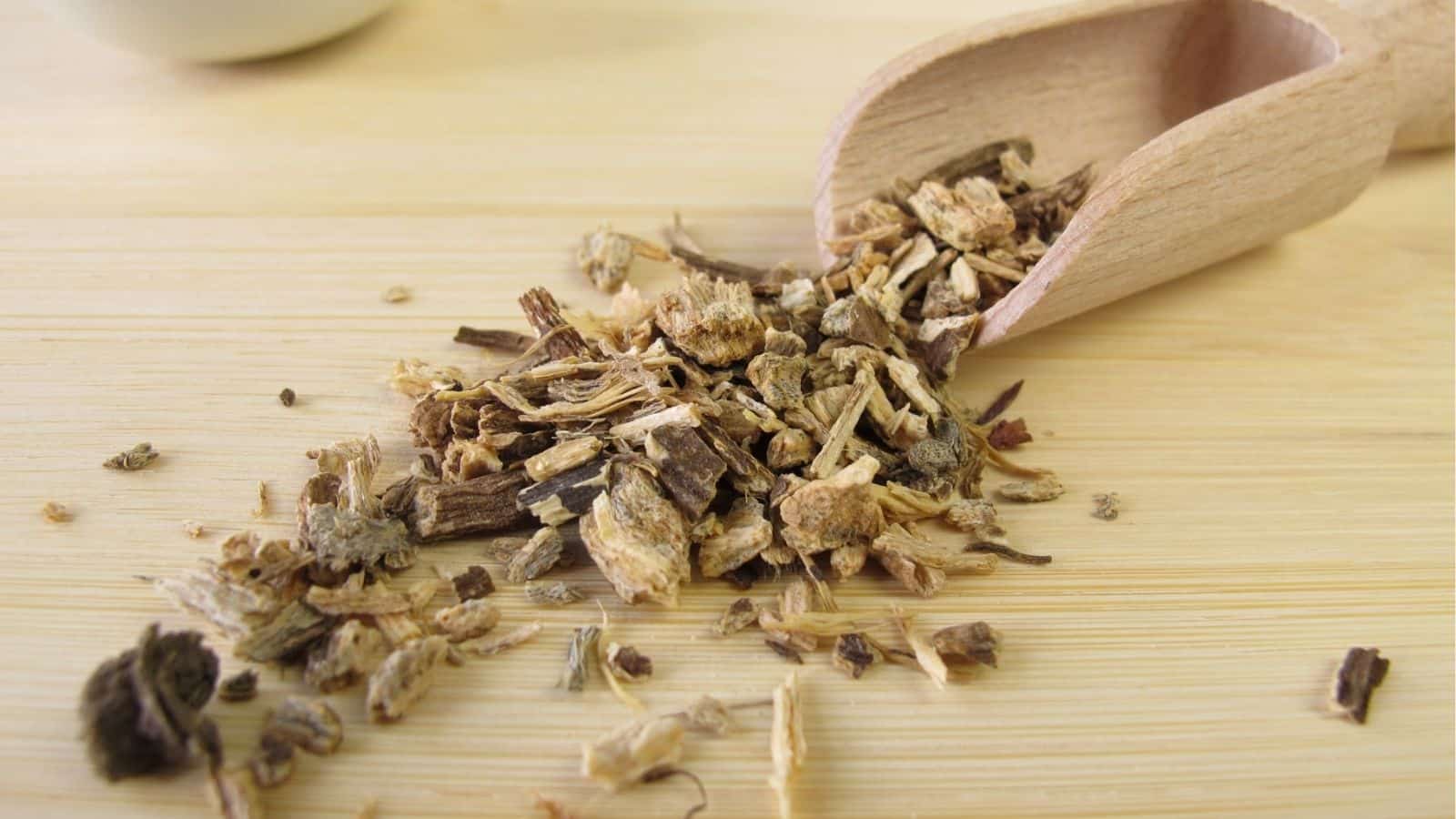A wooden scoop with dried chopped roots and herbal pieces spilling onto a light wooden surface.