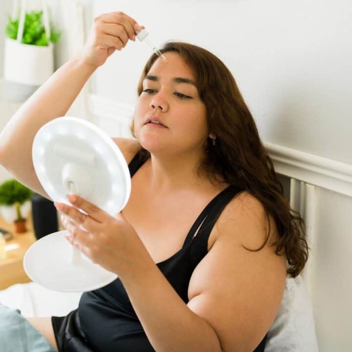 A woman sitting on a bed applies serum to her forehead using a dropper while holding a round mirror in her other hand.