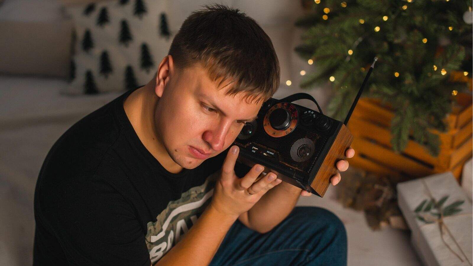 A man sits indoors holding a vintage radio to his ear next to a decorated Christmas tree and wrapped presents.