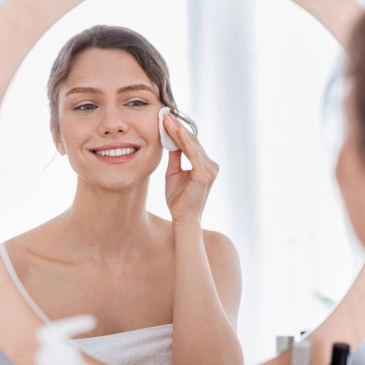 A woman wrapped in a towel applies a cotton pad to her face while looking in the bathroom mirror.
