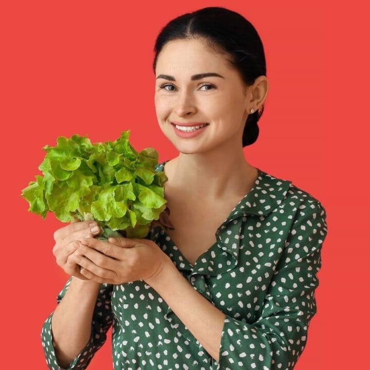 Woman holding a head of green lettuce and smiling, standing in front of a solid red background.