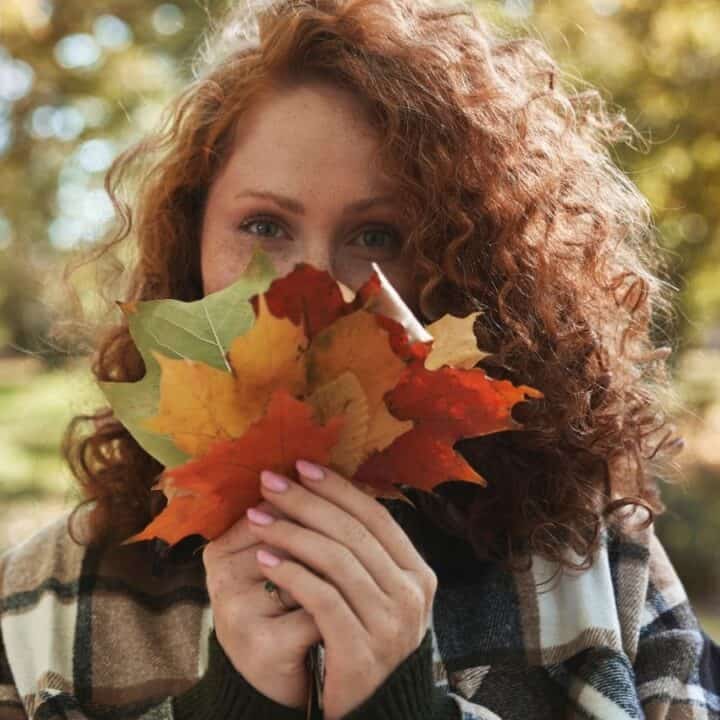A person with curly red hair holds a small bunch of autumn leaves in front of their face, outdoors in a park with trees in the background.
