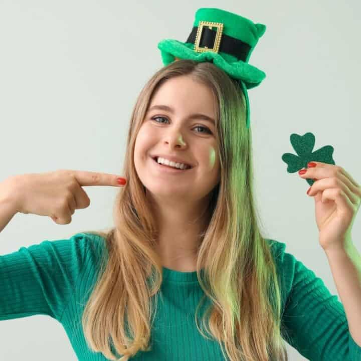Young woman in a green top and leprechaun hat smiles, pointing to herself and holding a green shamrock cutout against a plain background.
