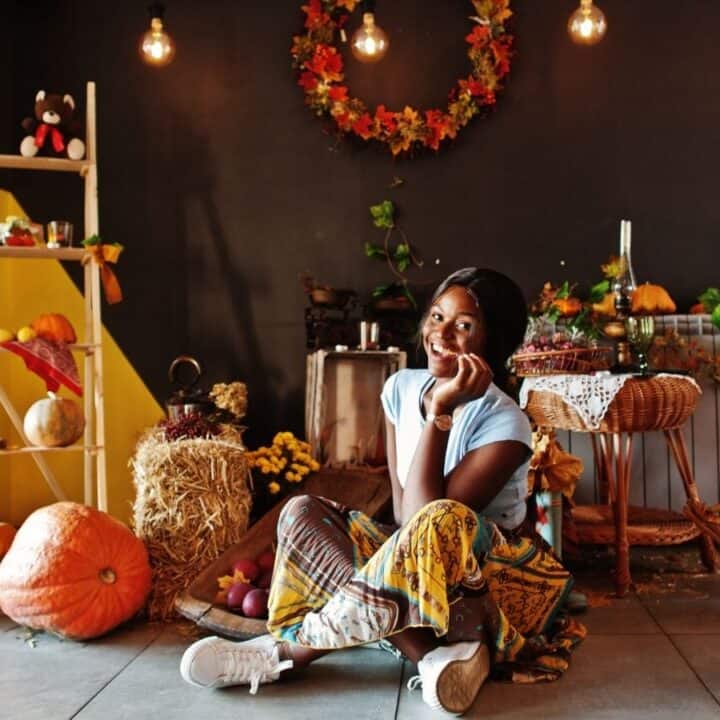 A woman sits on the floor in a room decorated with pumpkins, autumn leaves, candles, and a wicker chair, creating a cozy fall atmosphere.