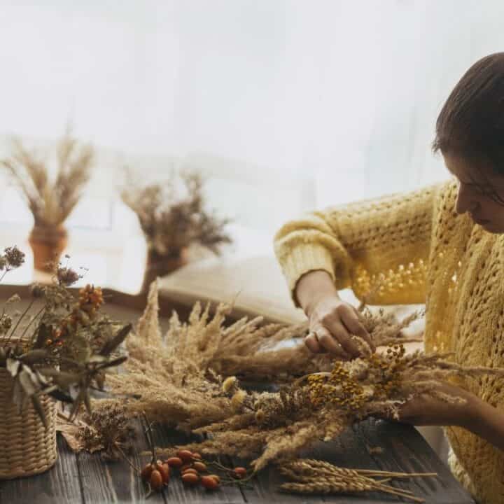 A woman in a yellow sweater arranges dried flowers and plants on a wooden table near a window with natural light.
