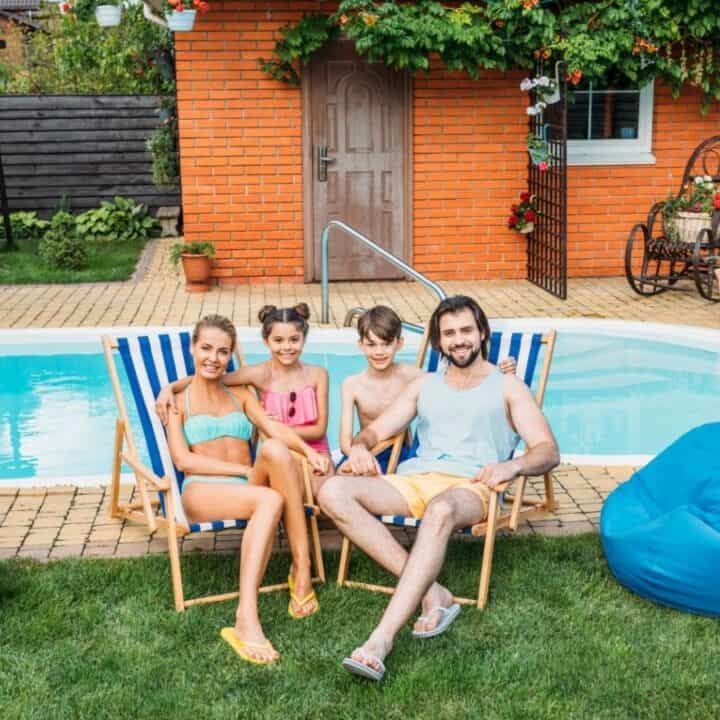 A family of four sits on deck chairs and smiles beside a backyard pool, with bean bags, a beach ball, and a brick house in the background.