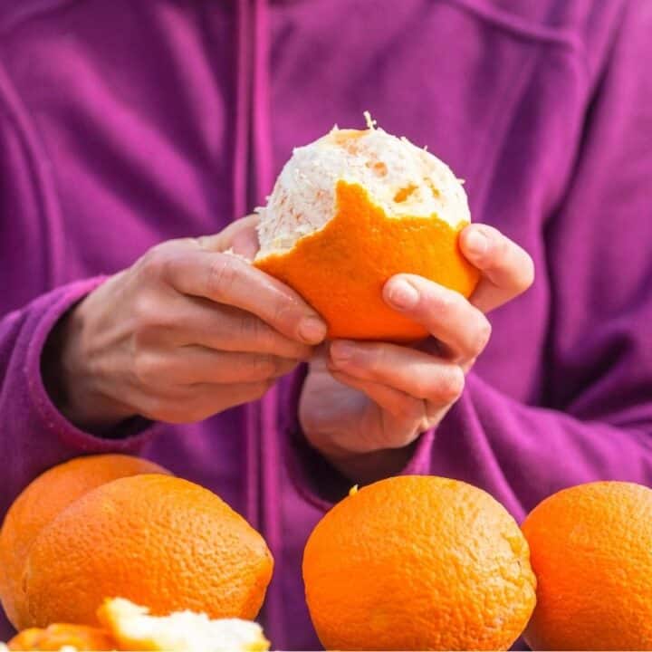 A person in a purple jacket peels an orange with several whole oranges on the table in front of them.