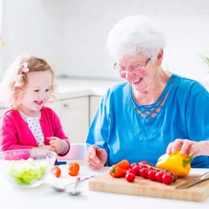 An elderly woman and a young girl sit at a kitchen table, smiling while preparing vegetables together.