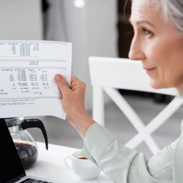 A woman sitting at a table holds up a paper document with printed financial information next to a laptop, a coffee pot, and a cup.