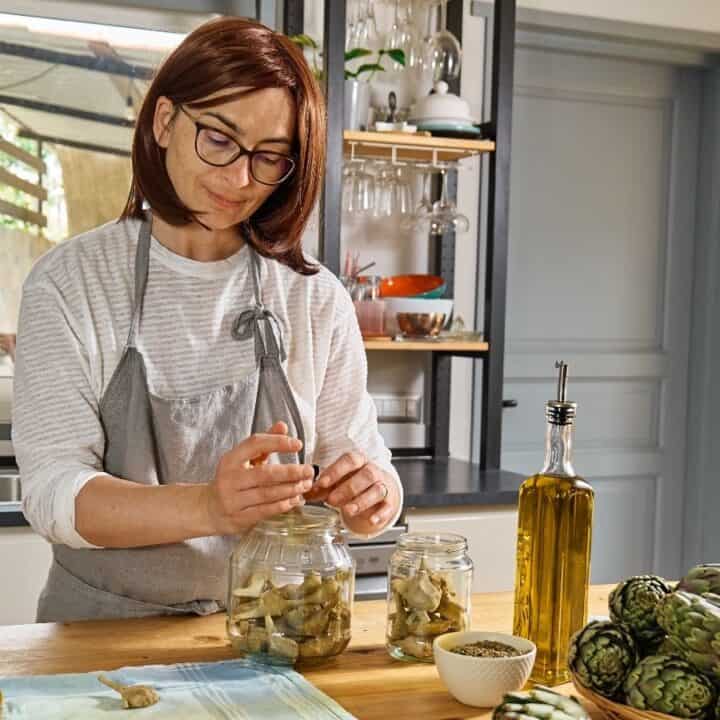 A woman in a kitchen places artichoke pieces into a glass jar. Artichokes, olive oil, and a jar of seeds are on the countertop.