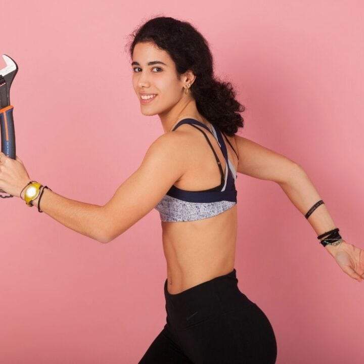 A woman in athletic wear holds a large adjustable wrench and poses as if running, against a pink background.