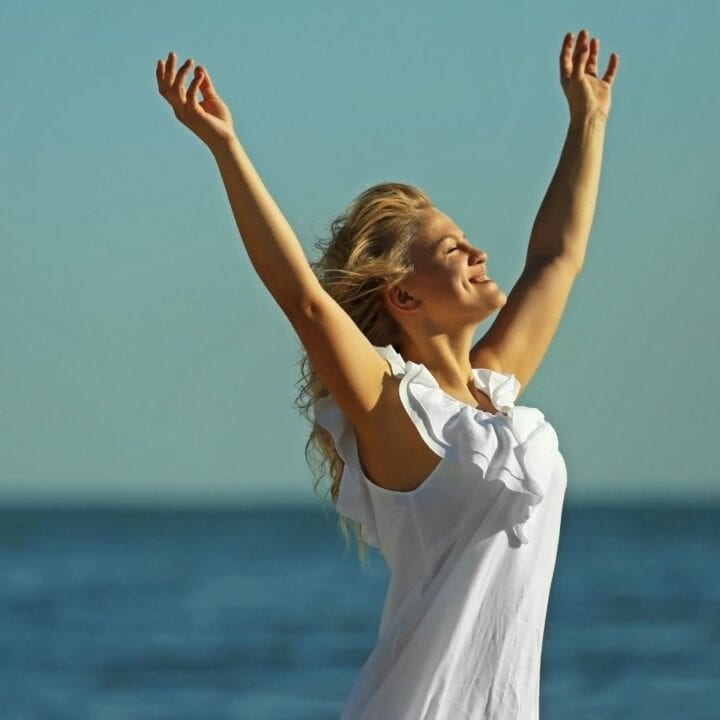 Woman in a white dress stands near the sea with arms raised, smiling and looking upward on a clear, sunny day.