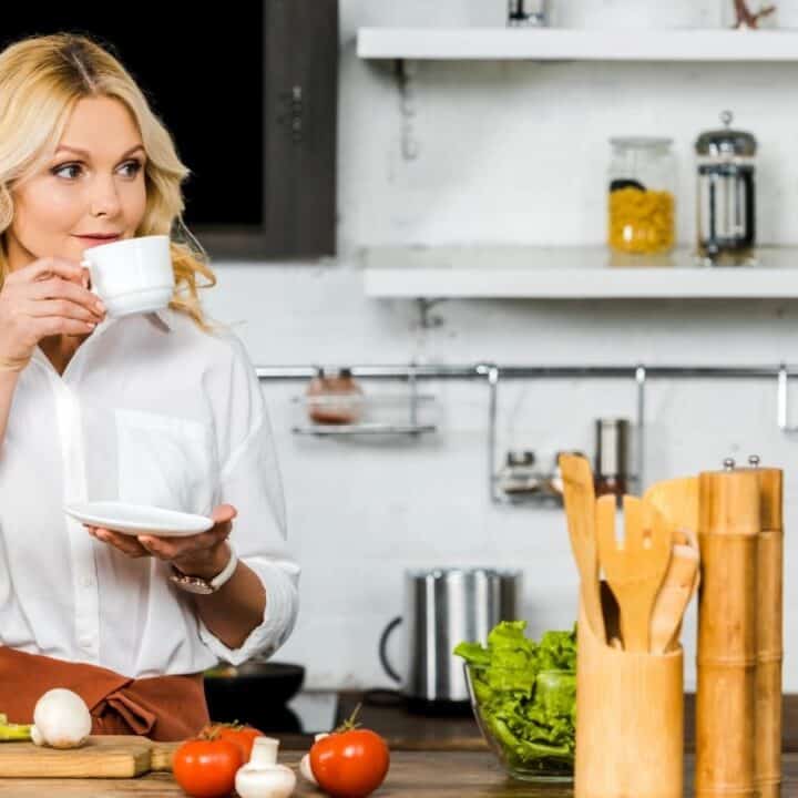 A woman in a white shirt stands in a kitchen, holding a cup and saucer, with vegetables and kitchen utensils on the counter in front of her.