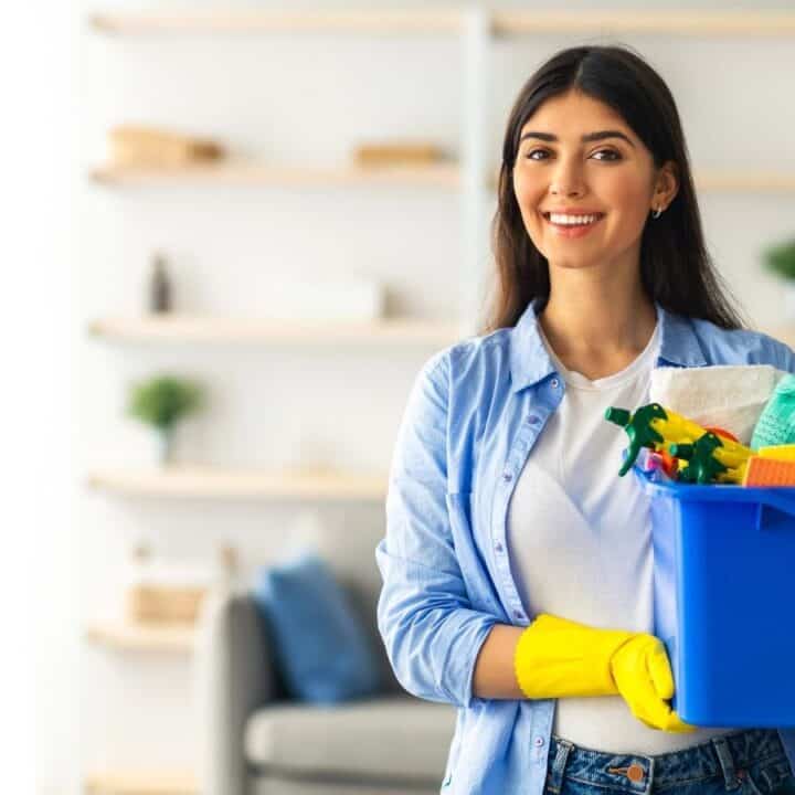 A woman wearing yellow cleaning gloves holds a blue bucket filled with cleaning supplies, standing in a living room with shelves in the background.