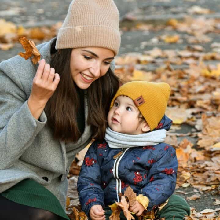 Woman and toddler sit on the ground covered with autumn leaves; the woman holds a leaf and they are both dressed in warm coats and hats.