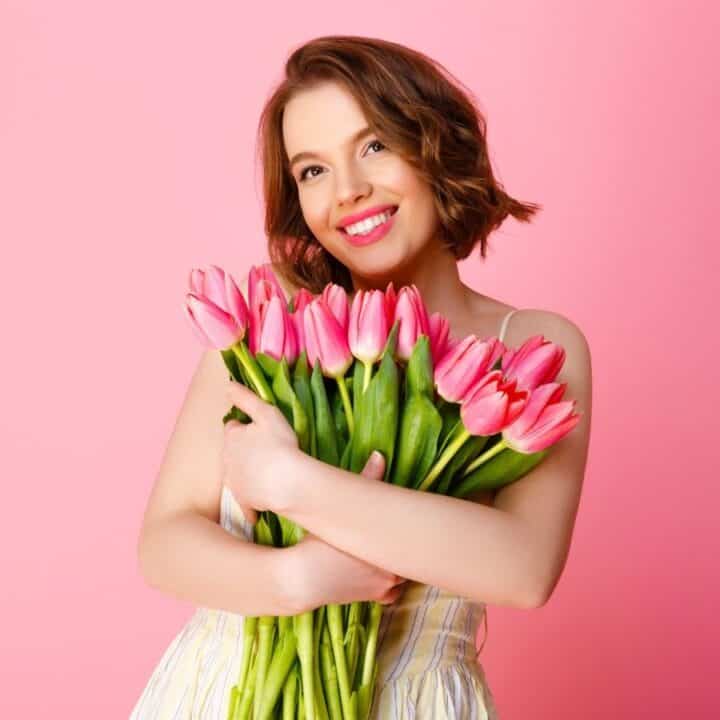 Woman in a light dress holding a bouquet of pink tulips, standing against a plain pink background, smiling at the camera.