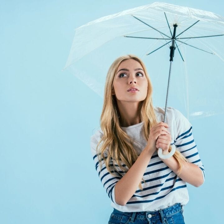 A woman with long blonde hair wearing a striped shirt holds a transparent umbrella, looking upwards against a light blue background.