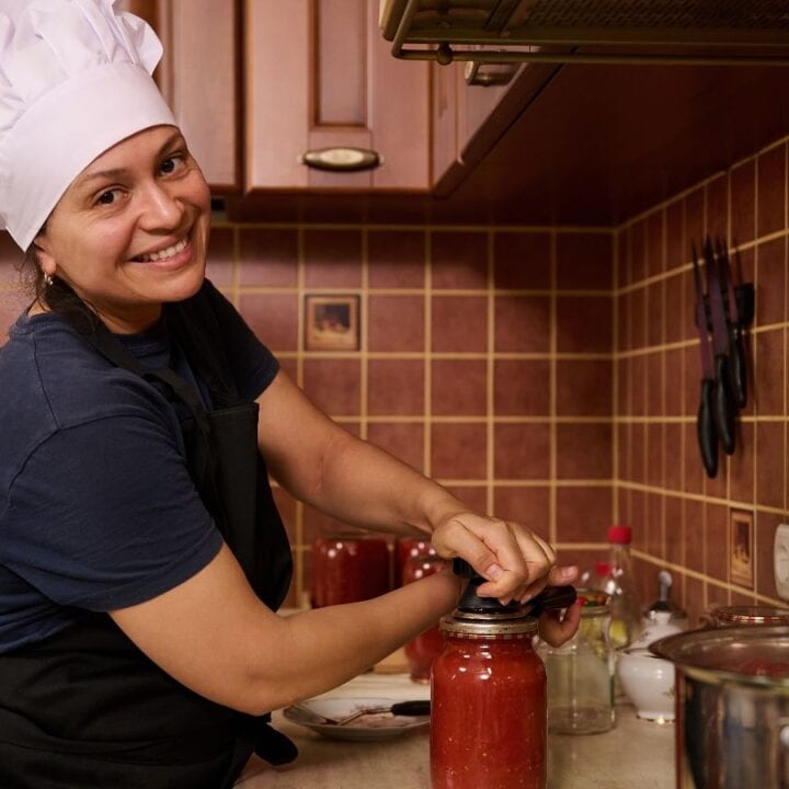 A woman in a chef hat and apron smiles while sealing a jar of tomato sauce in a kitchen with brown tiled walls and wooden cabinets.