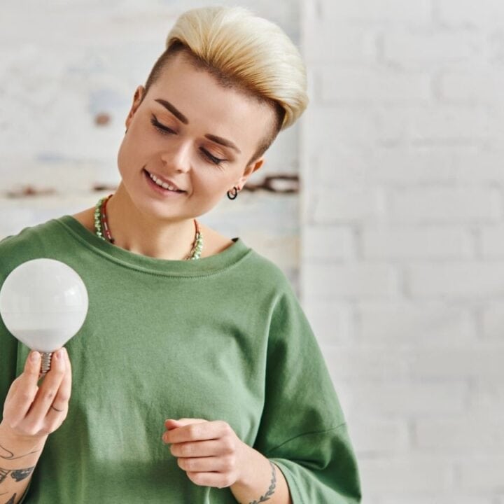 Person wearing a green shirt holds and looks at a large light bulb against a white brick wall background.